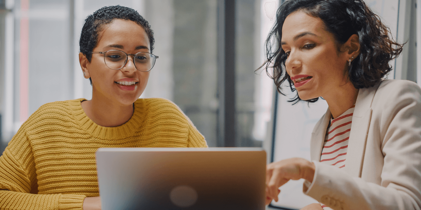 Two women of color collaborate around a laptop computer in a modern office space.