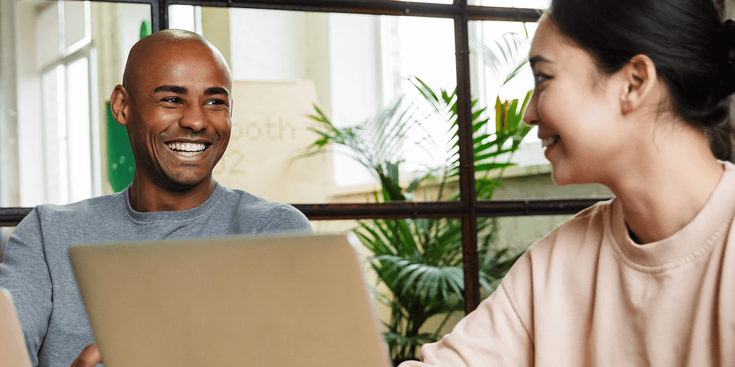 A Black man and an Asian woman collaborate sitting side-by-side with laptops. They are smiling and laughing.