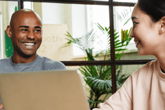 A Black man and an Asian woman collaborate sitting side-by-side with laptops. They are smiling and laughing.