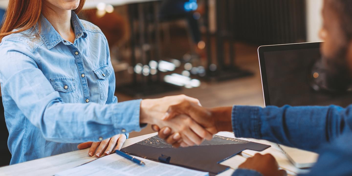 A white woman with red hair shakes hands across a desk with a Black man.