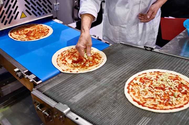 A food worker places shredded cheese onto an uncooked pizza crust with sauce on a food-safe conveyor belt.