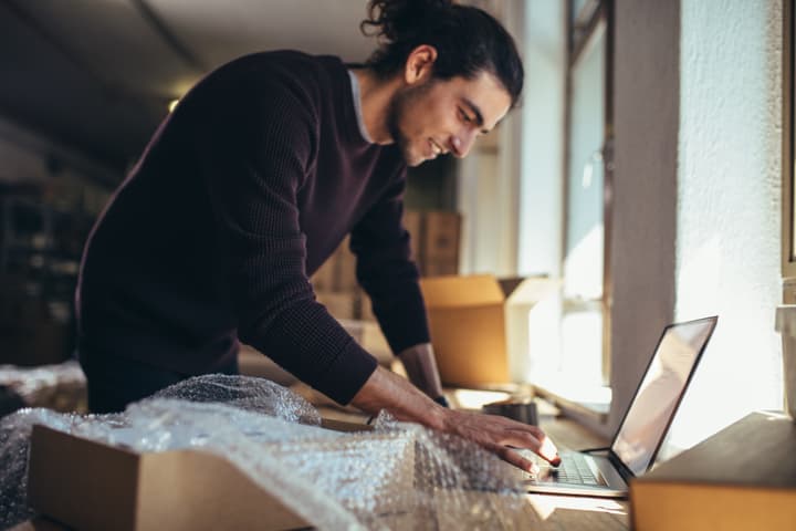 A man in a dark sweater with long, dark hair tied back into a bun updates the delivery status of a shipment on a laptop while inside a storage facility.