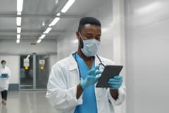 A Black male doctor wearing protective mask and gloves using tablet working in medical clinic.