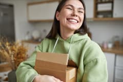 A woman in a light green hoodie hugs an opened shipping box while smiling wide, clearly pleased with the contents inside.