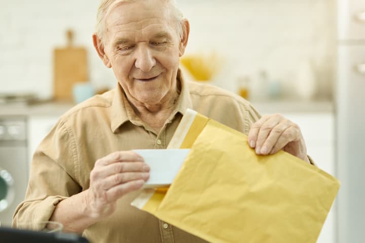 An elderly white man opens a padded mailer envelope to retrieve a small box of medication.