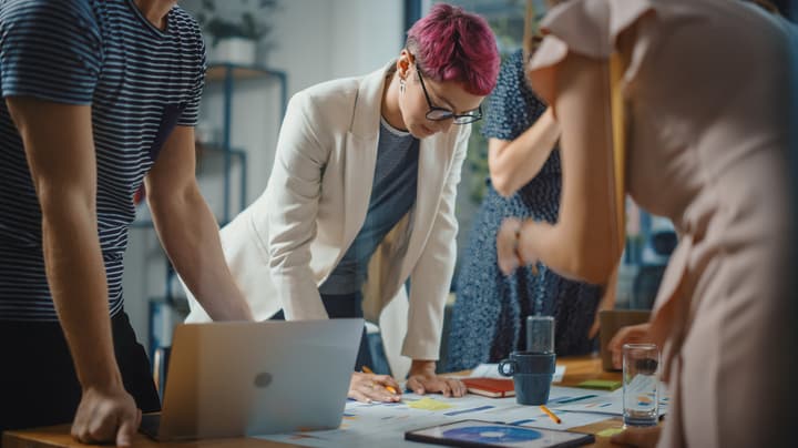 A woman with pink hair and glasses looks down at papers on a table with colleauges standing around the table.