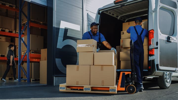 Two workers load a van with cardboard boxes outside of a logistics warehouse.