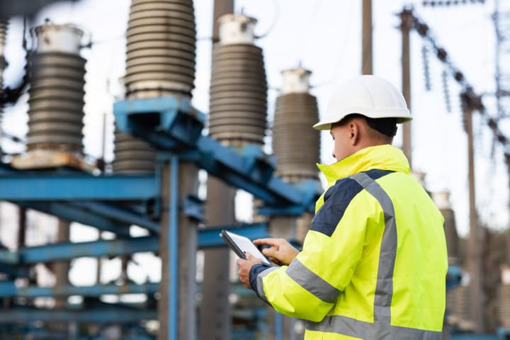 A white male field electrician records information about a power substation on a digital tablet. He is wearing a yellow, high visibility jacket and a white hard hat.