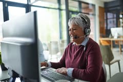 An older woman of color wearing a microphone headset takes a call from a customer while seated in front of a computer.