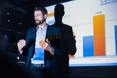 White male in business attire presents data from a projector to his colleagues.