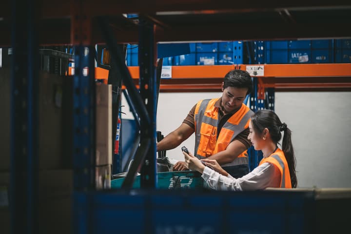 Two warehouse workers looking at a tablet with a forklift