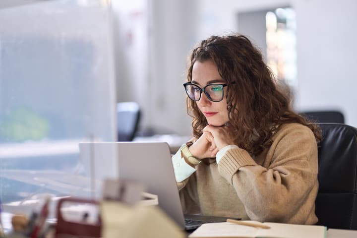 A woman with glasses looks intently at her laptop in an office.