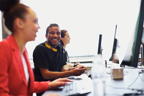 Three young business people of various races and genders work at computers in an open concept office. Focus is on the Black man who is candidly smiling towards the camera. He wears a pair of yellow headphones around his neck.