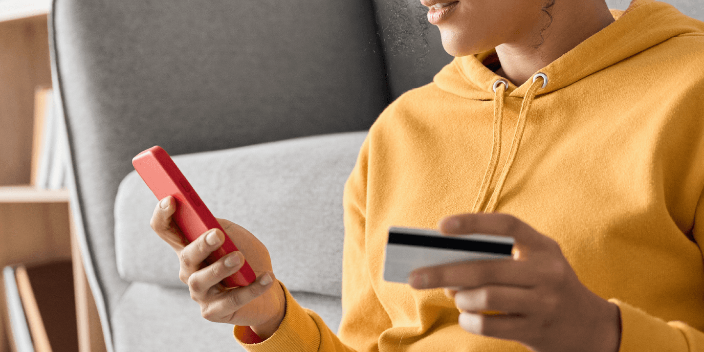 Close up of a person wearing a yellow hoodie holding a credit card in one hand and their smart phone in the other hand, implying they're making an online purchase through their mobile phone.