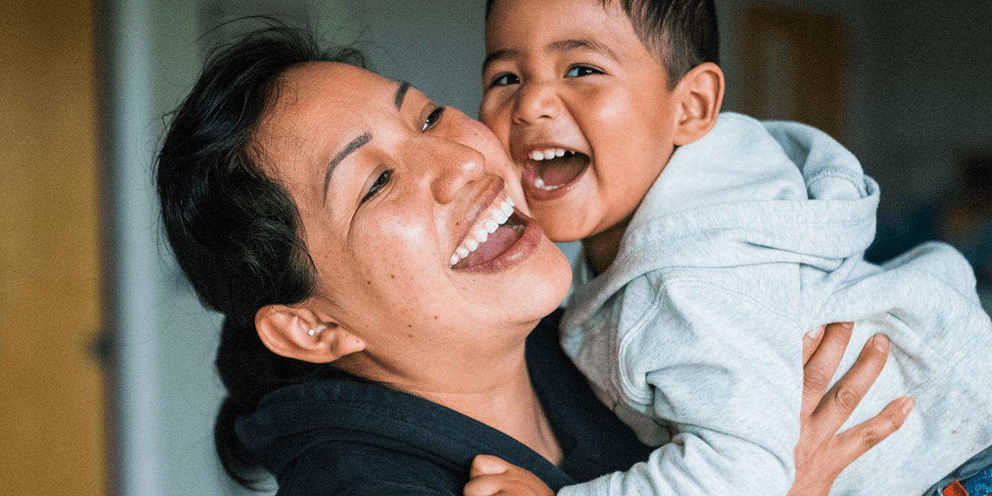 Woman of color holds up a young child of color – the child appears to be between 3 and 5 years old. They are both laughing and smiling.