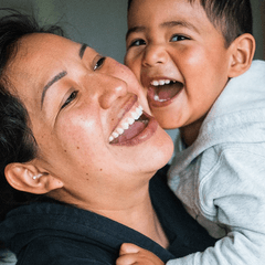 Woman of color holds up a young child of color – the child appears to be between 3 and 5 years old. They are both laughing and smiling.