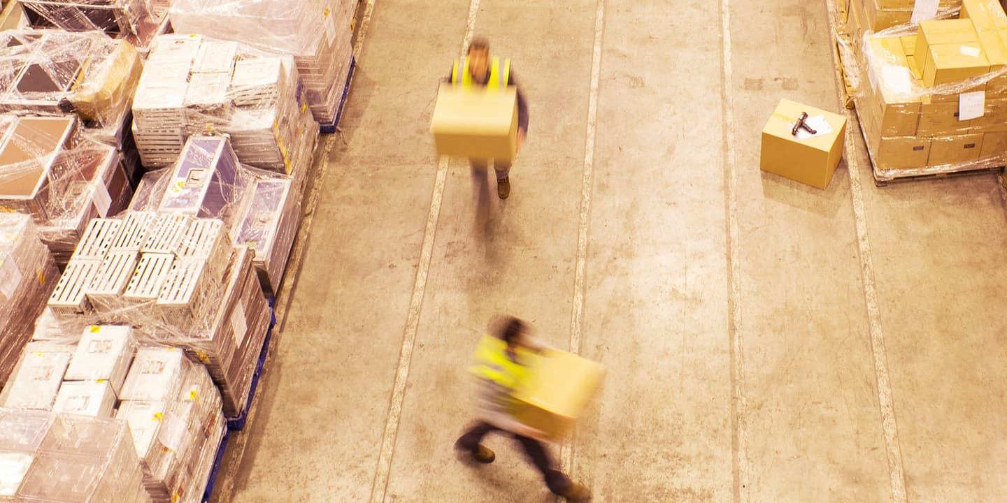 Overhead photograph of two workers in yellow, hi-vis vests moving large, cardboard boxes around a stock warehouse. They are moving so quickly both figures are blurred.