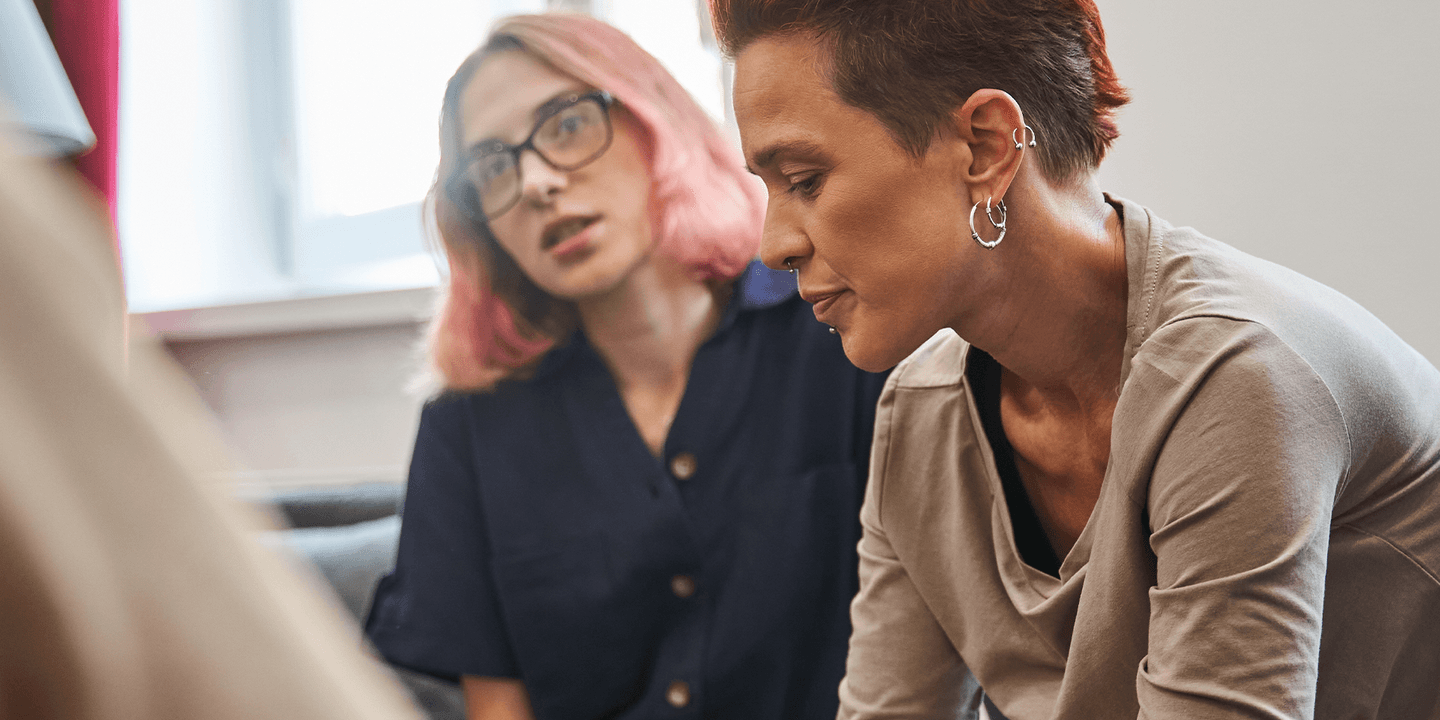 Two queer people solemnly share a moment. The person in the background seems to be comforting the person in the foreground. The shoulder of a third participant in the convo is close to the camera, but the rest of the person is located off-screen.