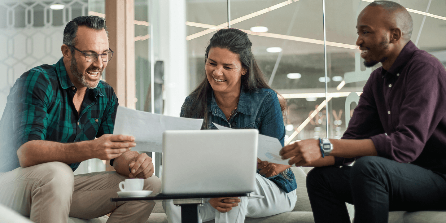Image of three business people: the left-most is an older white man, the middle is a woman of color, the right-most is a Black man. They are gathered around a small coffee table with a laptop on it. The white man and Black man are comparing printed pages while the woman of color joins the conversation.