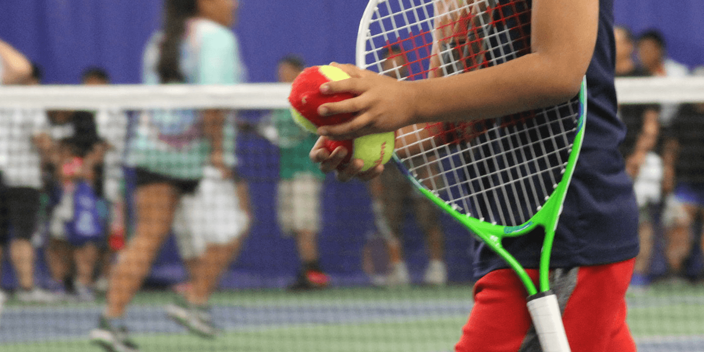 Close up of a child of color holding a tennis racket under their arm and carrying tennis balls in both hands.