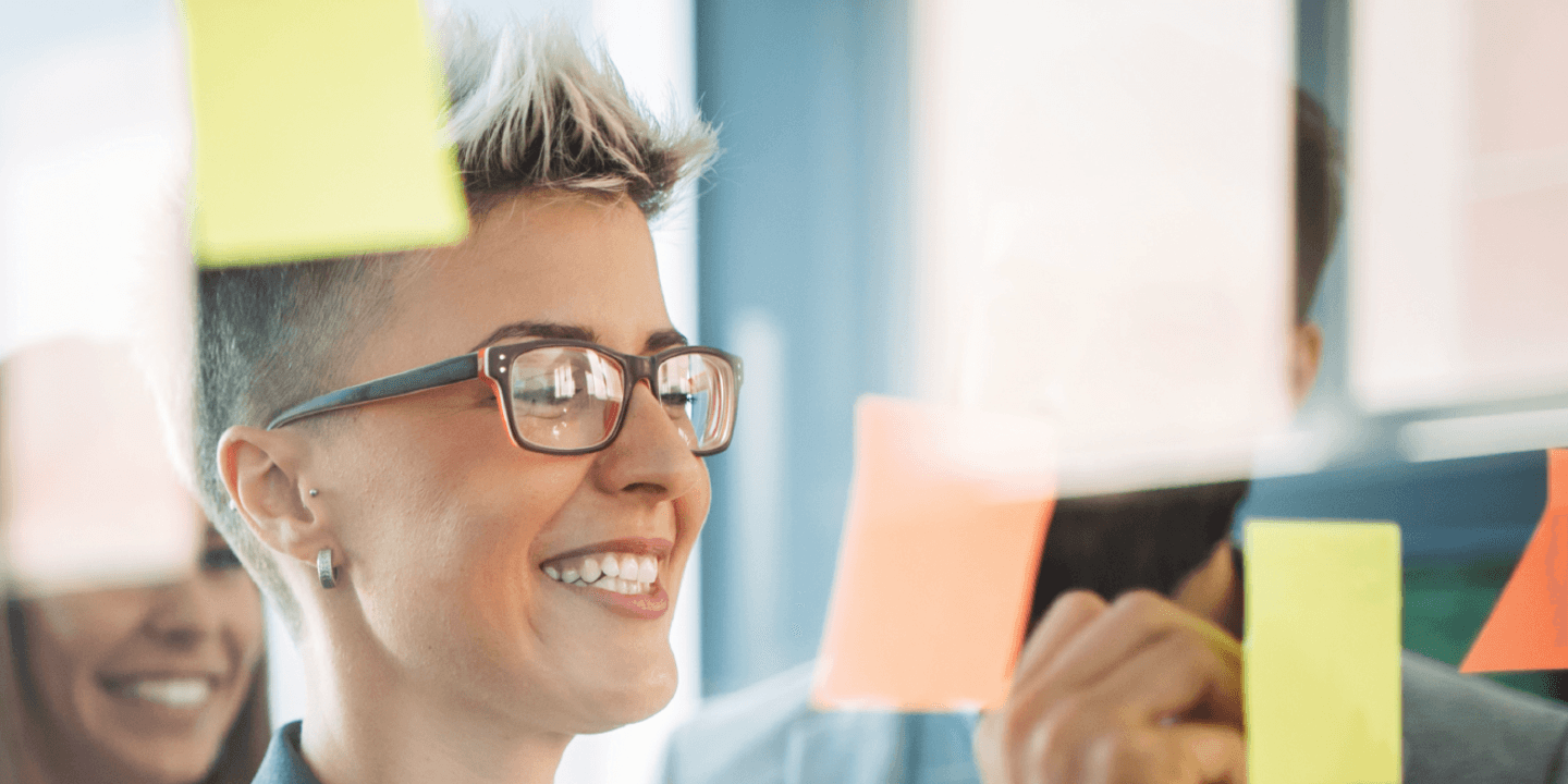 Image of business people working on a glass office partition, placing sticky notes on the glass for a project they're working on. They appear to be happy.