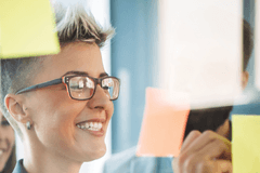Image of business people working on a glass office partition, placing sticky notes on the glass for a project they're working on. They appear to be happy.