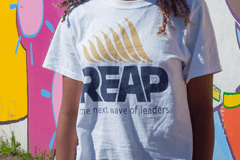 Close up of a young Black girl wearing a REAP t-shirt in front of an outdoor wall that is painted with a colorful mural.
