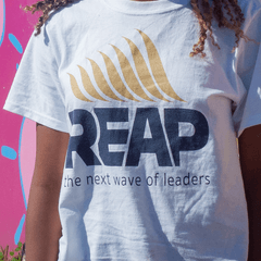 Close up of a young Black girl wearing a REAP t-shirt in front of an outdoor wall that is painted with a colorful mural.