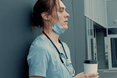 Image of a white, female medical professional wearing pale blue scrubs and a stethoscope. Her face mask is pulled down below her chin and she is holding a to-go cup of coffee. She appears exhausted.