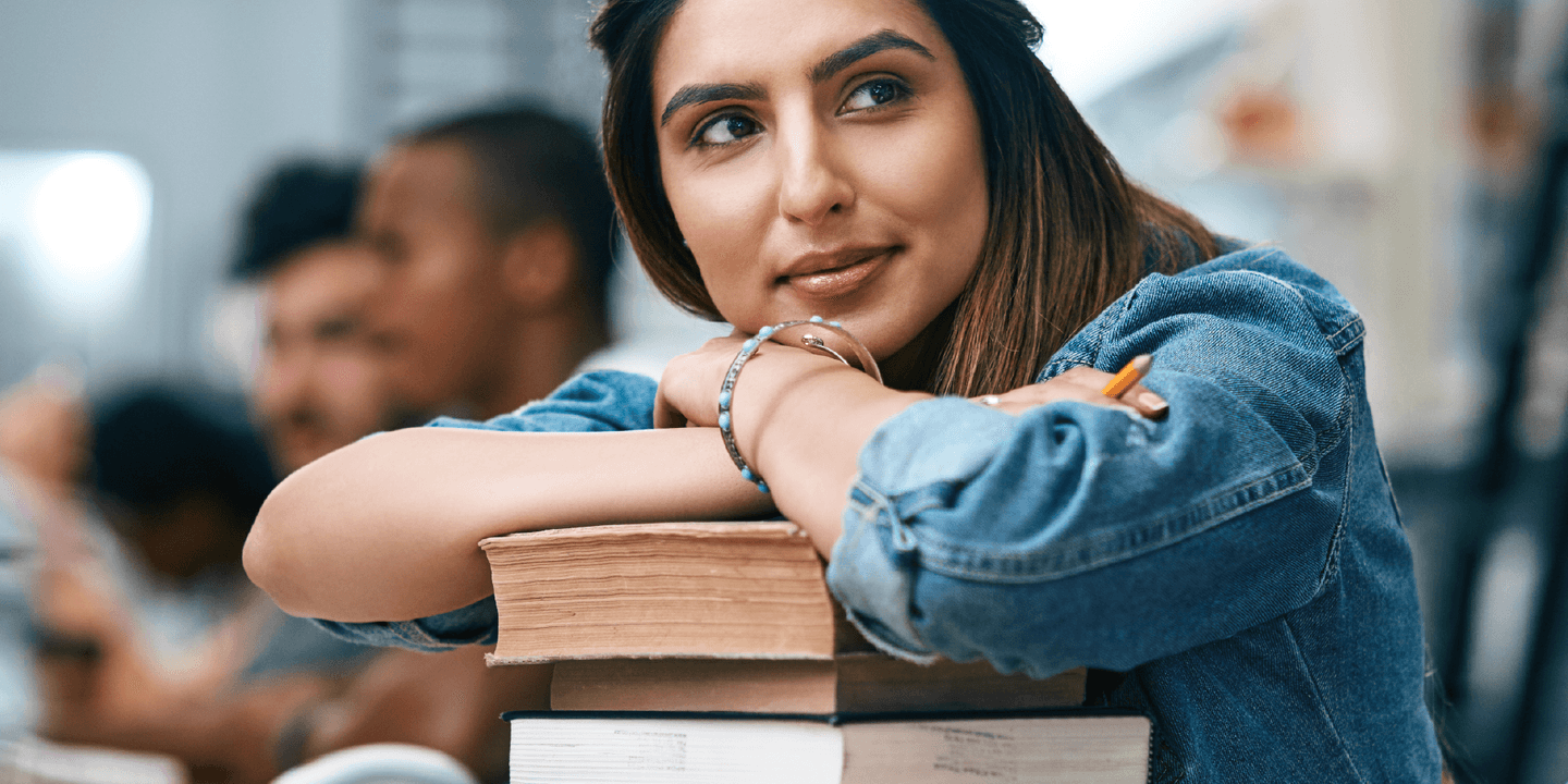 A light-skinned woman with dark hair and a denim shirt leaning on a pile of books with a slight smile on her face.