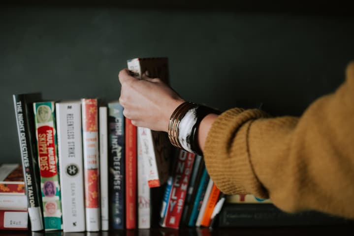 The arm of a white woman in a mustard sweater with metal bracelets grabs a book off a shelf of books. Photo by Christin Hume via unsplash.