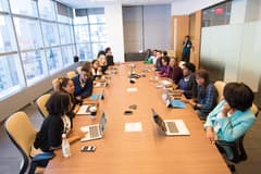 Over 15 people of color of various ages and genders gather around a large meeting table inside a modern office. Most attendees have brought a laptop. Photo by Christina Morillo on behalf of wocintechchat.com via Unsplash.