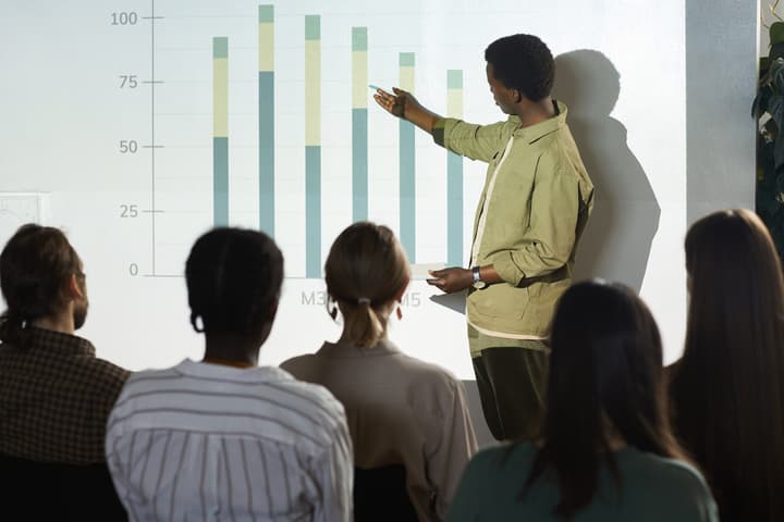 Portrait of young Black man pointing at data graph while giving speech or report on statistics during business conference to a group of people of various ages, races, and genders.
