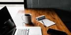 A wooden desk with the following resting on its surface: a MacBook laptop, a memo pad with illegible writing, a white mug full of black coffee, and a smart phone. Photo taken by Andrew Neel via Unsplash