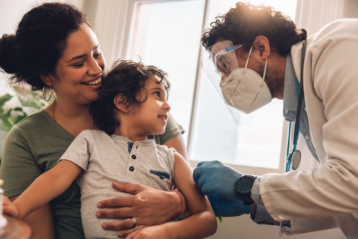 Doctor with a face mask on and gloves Caring For Toddler Sitting On Mother's Lap