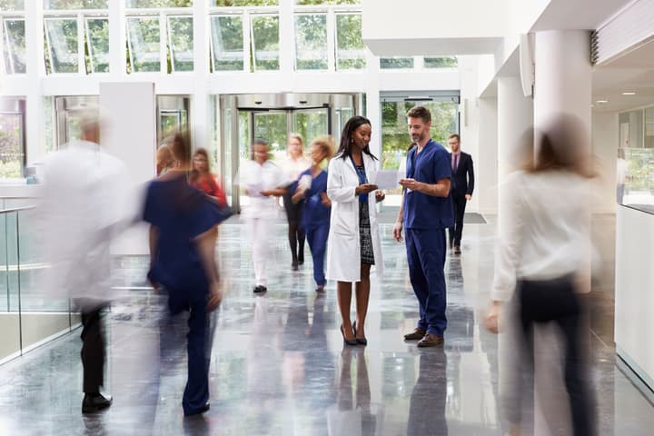 Staff In Busy Lobby Area Of Modern Hospital. Female doctor talking to male nurse with the blurred motion of people walking near them.