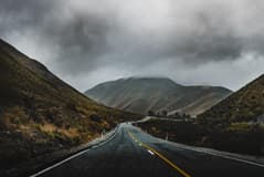 A paved, two lane road winding through desert hills on a gloomy, overcast day. Image by Iswanto Arif via Unsplash.