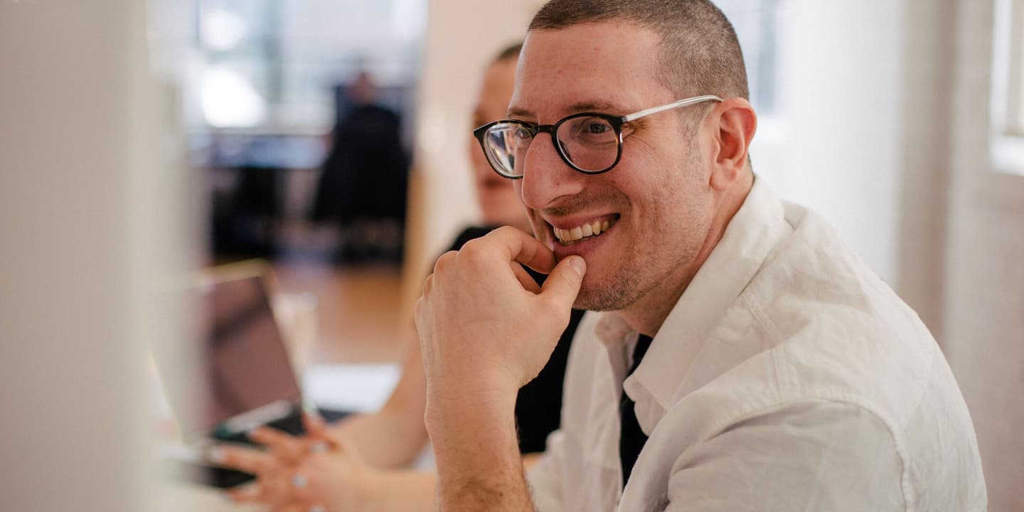 A millennial-aged white male smiles in a modern office setting. He appears to be engaged in a lively conversation with colleagues.