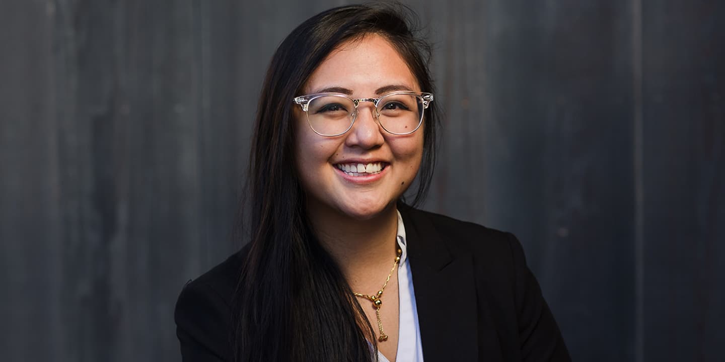 A portrait of Propeller employee Lena Chhay. She wears a white blouse and a black blazer in front of a dark, grey, wooden background. She wears glasses with a clear, acrylic frame.