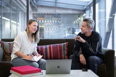 A white female and an older white male discuss a work-related topic while seated in a modern office setting with comfortable furniture. A laptop is on the coffee table in front of the couch they're sitting on. Photo by LinkedIn Sales Solutions via Unsplash.