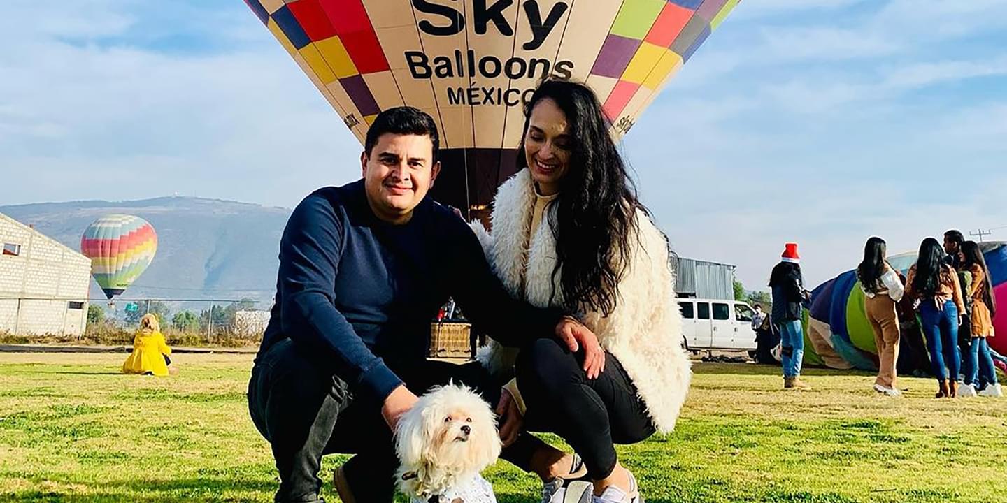 Image of Linda Villarreall, her husband, and her dog. They are posing outside on a sunny day in front of a colorful hot air balloon.