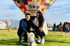 Image of Linda Villarreall, her husband, and her dog. They are posing outside on a sunny day in front of a colorful hot air balloon.