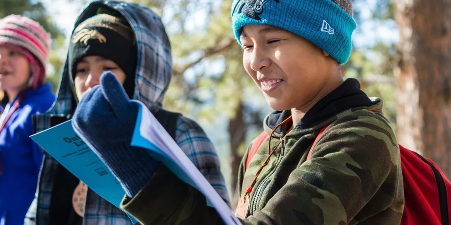Three children dressed warmly in beanies, gloves, and jackets, consult their Outdoor Lab Foundation guide book during a nature hike. They are smiling and happy.