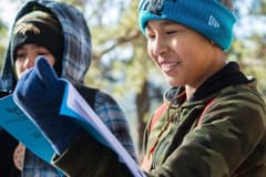 Three children dressed warmly in beanies, gloves, and jackets, consult their Outdoor Lab Foundation guide book during a nature hike. They are smiling and happy.