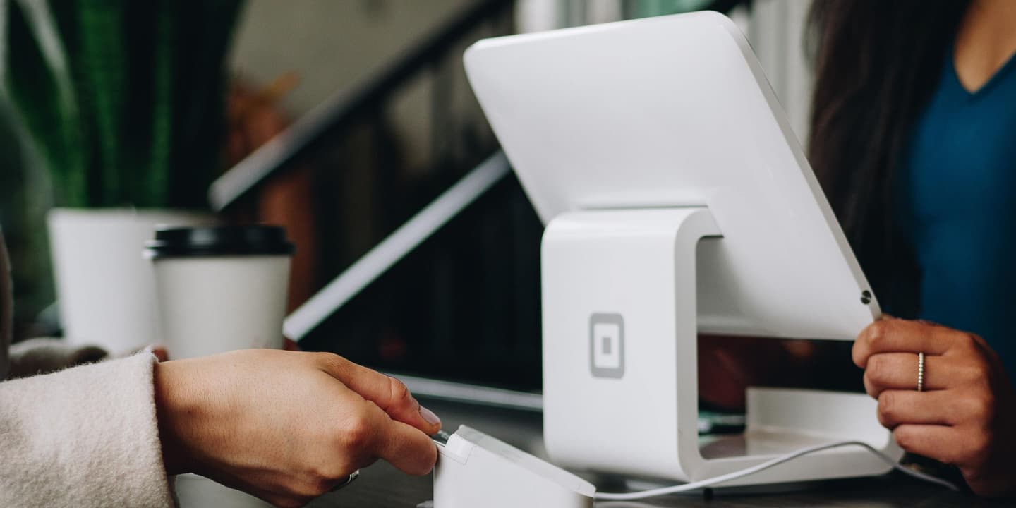 Close up of a feminine hand paying for their purchase with a Square card reader at a Square point of sale system; the cashier is a Black woman. Photo by Patrick Tomasso via Unsplash.