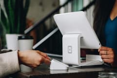 Close up of a feminine hand paying for their purchase with a Square card reader at a Square point of sale system; the cashier is a Black woman. Photo by Patrick Tomasso via Unsplash.