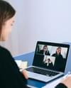 A white woman in a black blazer sitting in front of a laptop talking to three people on a video call while taking notes on a piece of paper. Photo by Anna Shvets via pexels.