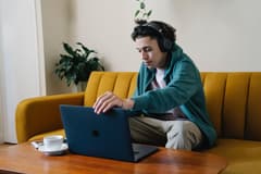 Serious man of color in wireless headphones using a laptop on a coffee table in front of a couch. It appears he is working remotely. Photo by Eren Li via Unsplash.