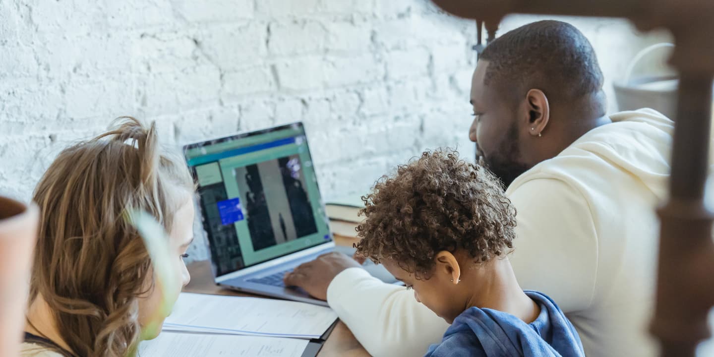 A image of a Black father working on a laptop from home while his wife feed their infant child, and their young child stands near his father's desk. Photo by Keira Burton via Unsplash.