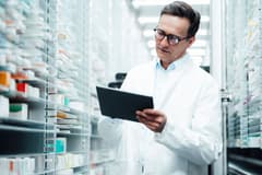 An older, white male pharmacist consults a digital tablet while surrounded by cases with dozens of shelves of medications.